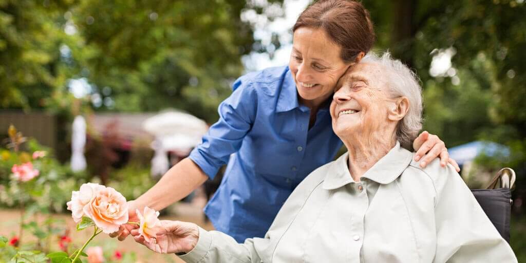 A disabled senior woman in a wheelchair touching flowers with the help of a younger woman caregiver.