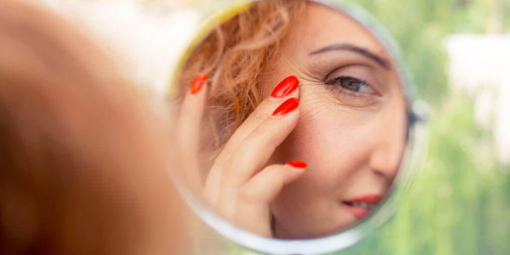 A middle-aged woman is touching her face while looking in a mirror.